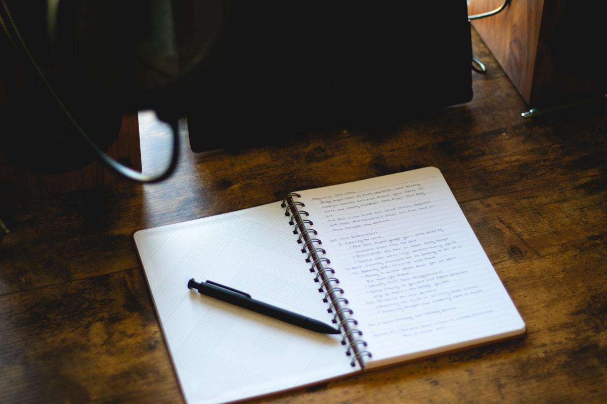 An open notebook with a pen resting on it, on a plain wooden desk