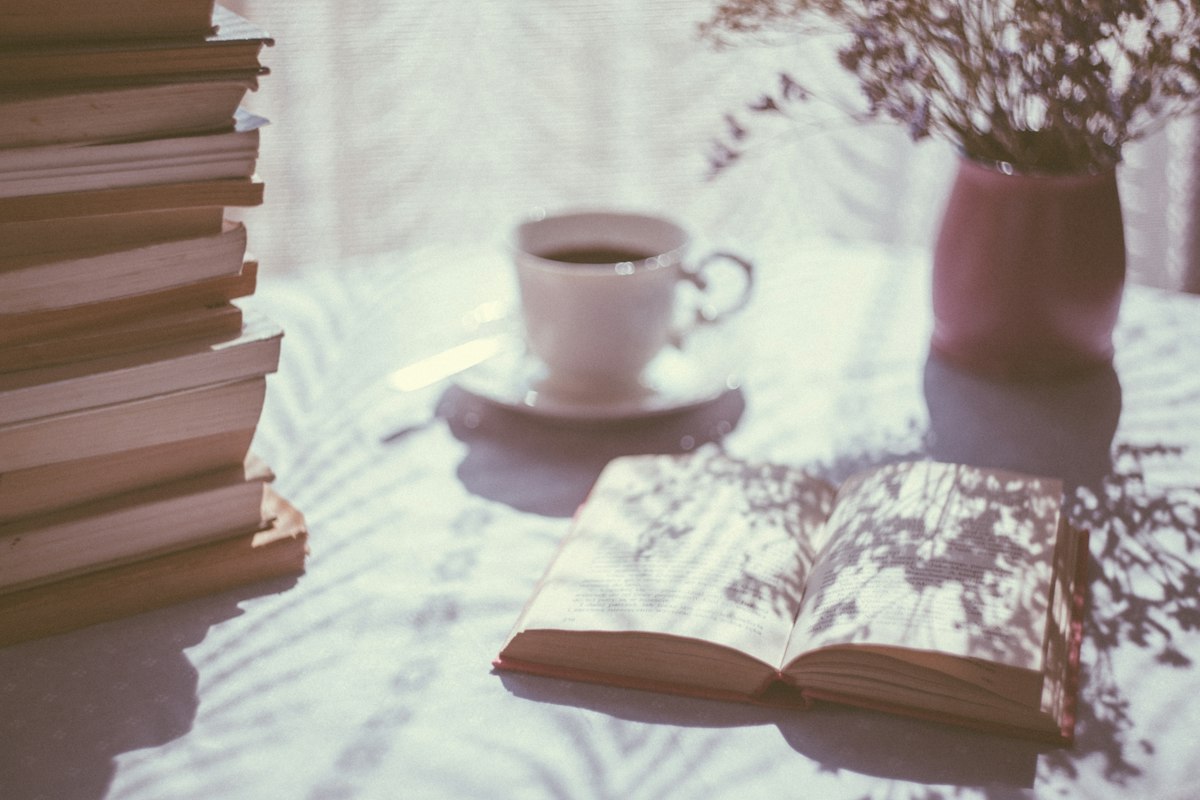 An open book and a cup of coffee on a sunlit table beside a stack of books