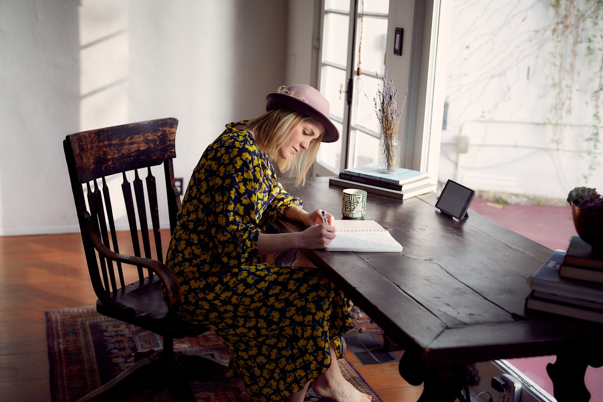 A person writing thoughtfully in a journal by a window, warm light falling across the open page